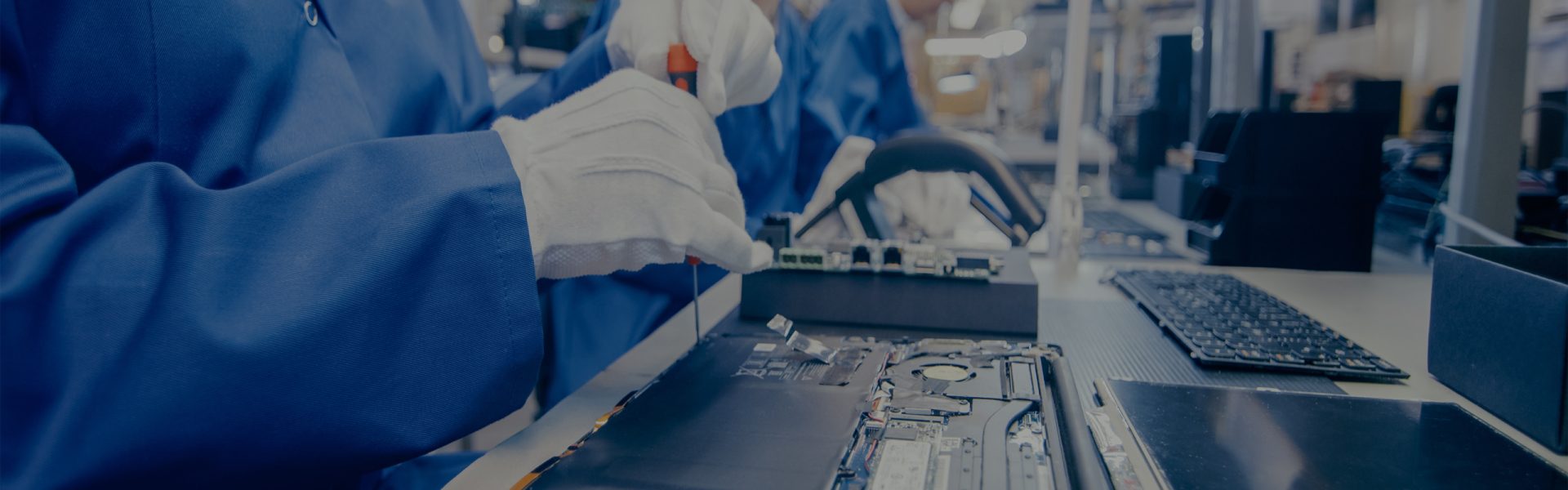 Close-Up of a Female Electronics Factory Worker in Blue Work Coat Assembling Laptop's Motherboard with a Screwdriver. High Tech Factory Facility with Multiple Employees.